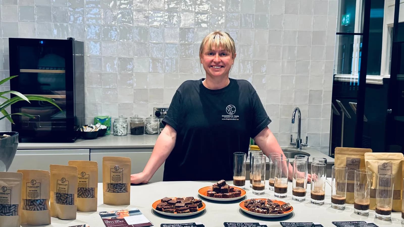 Niki Charnock behind a table set out for a wellness talk, with pouches of Gut Buster herbal capsules, plates of raw flapjack and small glasses of drink