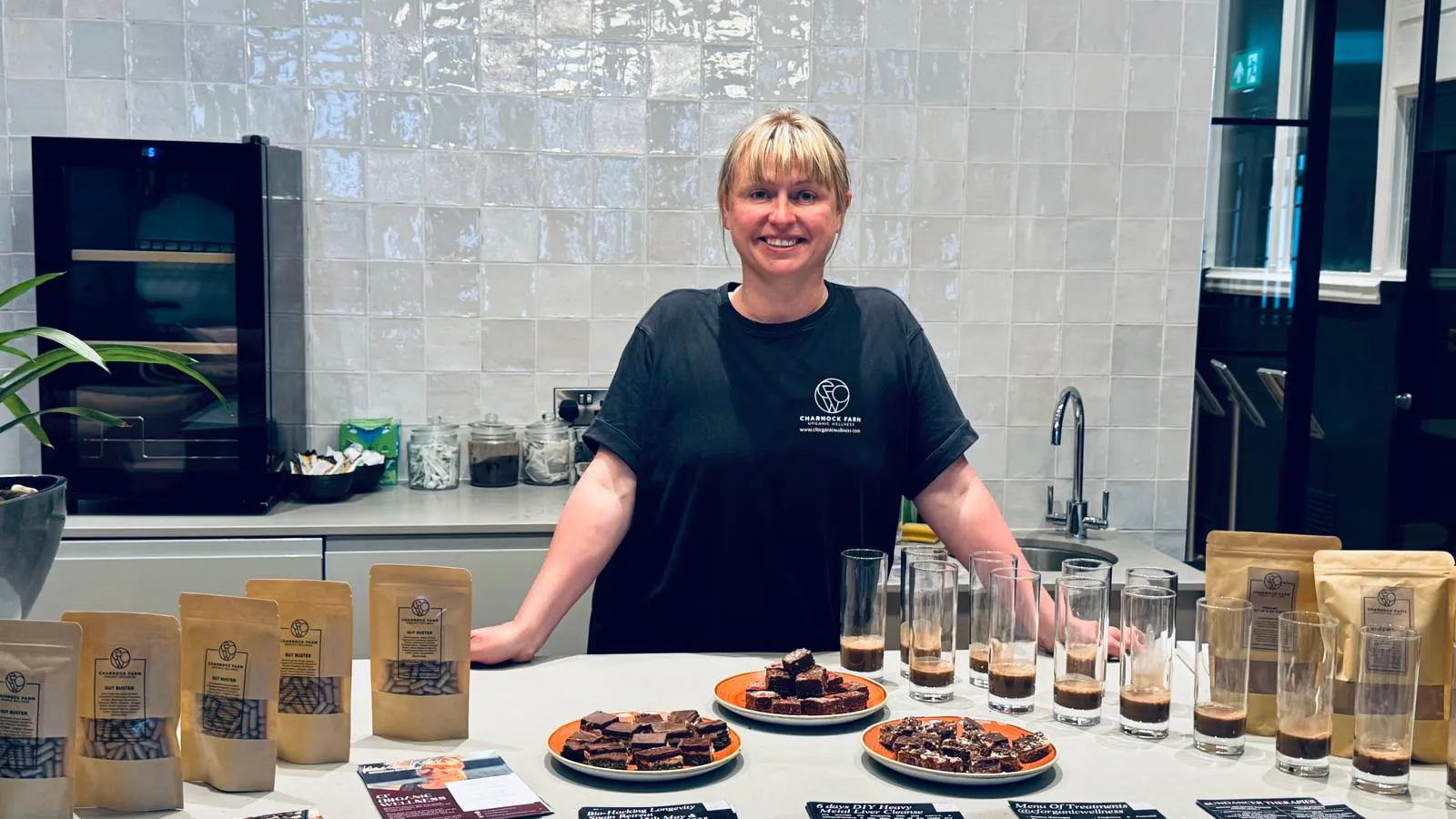 Niki Charnock behind a table set out for a wellness talk, with pouches of Gut Buster herbal capsules, plates of raw flapjack and small glasses of drink